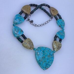 May include: A turquoise and tan stone necklace with a dark chain and clasp. The necklace features large, teardrop-shaped turquoise stones and smaller tan stones, connected by dark beads. The necklace is displayed on a white surface.
