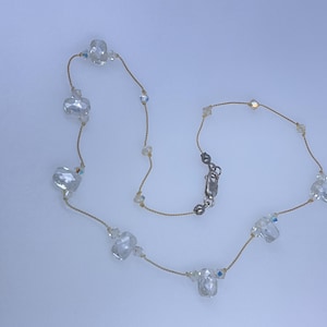 May include: A delicate necklace featuring clear, faceted crystal beads of varying sizes strung on a thin, gold-colored cord. The necklace has a silver-colored clasp and small, clear accent beads. The crystals reflect light, creating a sparkling effect.