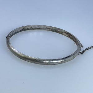 May include: A silver bangle bracelet with a delicate floral design and a small chain attached. The bracelet is marked "925" on the inside.