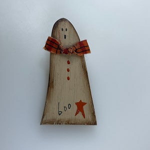 May include: A wooden ghost figurine with a red and orange plaid scarf, three red dots, and the word "BOO" written on the bottom. A red star is painted on the right side of the ghost.