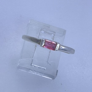 May include: A silver ring with a rectangular, pink gemstone. The ring is set on a clear display stand against a white background. The gemstone is a vibrant pink color, and the band is a simple, polished silver.