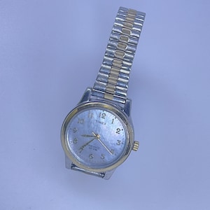 May include: A silver and gold-tone Timex watch with a light blue face. The watch has a silver band with gold accents and a gold-tone bezel. The watch face displays the numbers 1 through 12 and the Timex logo.