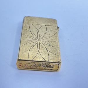 May include: A vintage gold-colored butane lighter with a floral design etched on the front. The lighter has a rectangular shape and the text "Scripto Butane U.S.A." is visible on the bottom.