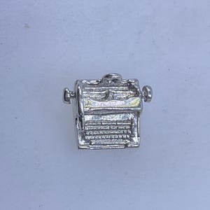 May include: A small, silver-colored typewriter charm. The charm has detailed keys, a carriage return lever, and side knobs. The typewriter is a miniature replica, likely for jewelry or crafting purposes. The charm is set against a plain, light background.