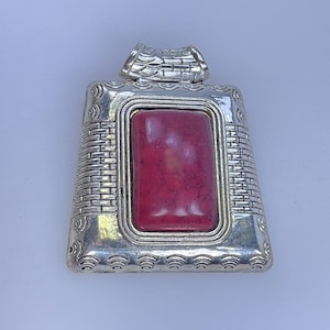 May include: Silver-toned rectangular pendant with a large, rectangular, pink-red stone center. The pendant has a decorative border with a brick-like pattern and a loop at the top for a chain. The overall design is ornate and eye-catching.
