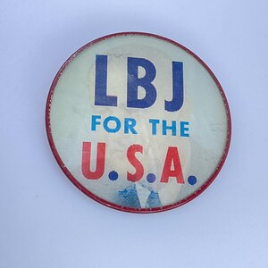 May include: Round button with a red border and the text "LBJ FOR THE U.S.A." in blue and red. The button has a light background with a faded image of a person.