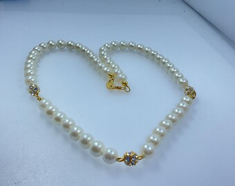 May include: A pearl necklace with a gold clasp and accents. The necklace features a strand of white, round pearls. Gold-colored metal settings hold small, clear crystals at intervals along the necklace.