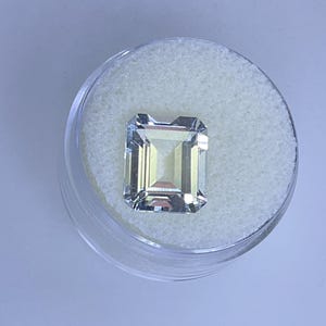 May include: A clear, emerald-cut gemstone displayed in a clear plastic container with a white, textured interior. The gemstone has a rectangular shape with a faceted surface, reflecting light. The container sits on a light blue surface.