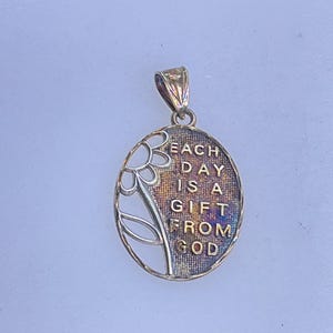 May include: Oval pendant with a floral design and the inscription "EACH DAY IS A GIFT FROM GOD." The pendant is silver-toned with a gold-colored bail. The background has a textured, iridescent finish.
