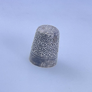 May include: A metallic thimble featuring a detailed, repeating geometric star pattern across its dome and a smooth, plain band encircling the base. The surface has a weathered, dark grey finish with visible signs of age and texture throughout.