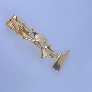 May include: A gold-colored tie clip with a detailed design. The clip features a stylized figure with a hat and a tool, possibly a representation of a profession or hobby. The clip is on a light blue background.