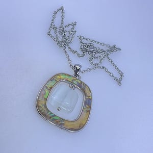 May include: Silver-toned necklace with a square pendant. The pendant features a white, rectangular center stone, framed by a silver border and an iridescent, abalone-like outer layer. The chain is delicate and hangs on a white background.