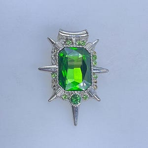 May include: A silver-toned brooch with a large, rectangular, emerald-green gemstone at its center. The brooch is adorned with smaller green gems and silver spikes radiating outwards, creating a starburst design. The brooch has a curved top.