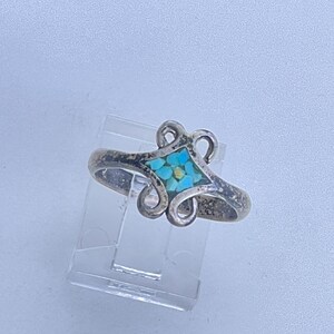 May include: A silver ring with a turquoise stone inlay in a floral design. The ring has a vintage, tarnished appearance and is displayed on a clear plastic stand. The turquoise is a bright blue color.