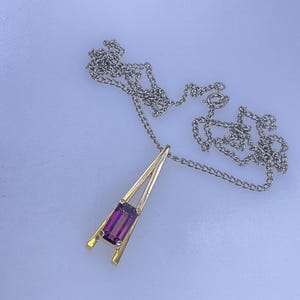 May include: A necklace featuring a silver-toned chain and a gold-toned pendant with a triangular frame. The pendant holds a single, rectangular, deep purple gemstone set vertically within the lower portion of the open metal structure.
