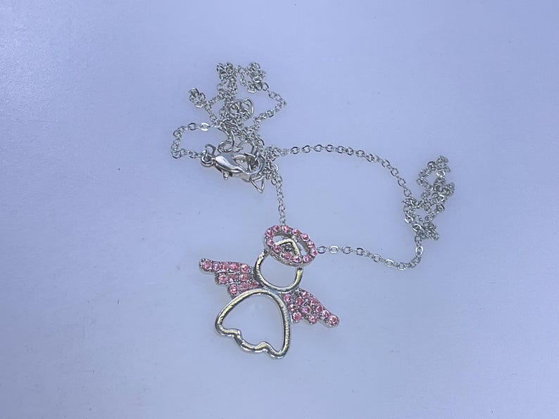 May include: A silver-colored necklace with a pendant shaped like an angel. The angel pendant has pink crystal accents on the wings and halo. The necklace chain is delicate and has a lobster clasp.