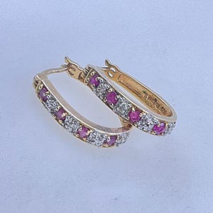 May include: A pair of gold hoop earrings with alternating red and clear gemstones. The earrings are oval-shaped and feature a hinged closure. The gemstones are set in a row along the front of the hoops.