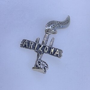 May include: A silver-toned charm featuring the word "ARIZONA" across a cactus design. Above the cactus is a banner that reads "GRAND CANYON ARIZ." The charm has a detailed, textured appearance and is likely a souvenir or travel-themed item.