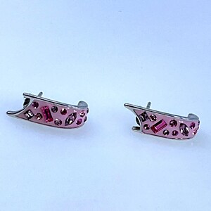 May include: A pair of pink and silver earrings. The earrings have a curved design with a pink enamel finish and are adorned with various sizes of pink and red crystals. The earrings are set against a white background.
