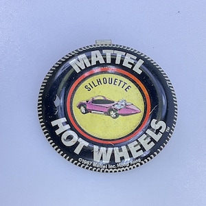 May include: A round, black Hot Wheels Silhouette button with a silver clasp. The center features a purple car on a yellow background, with the words "MATTEL SILHOUETTE HOT WHEELS" in white. The bottom reads "©1967 Mattel Inc. Hong Kong."