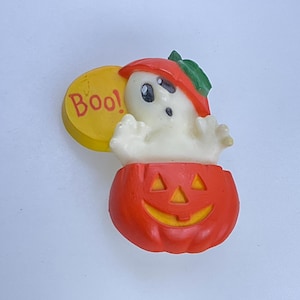May include: A Halloween-themed decoration featuring a white ghost wearing a red hat emerging from an orange pumpkin. A yellow circle with the word "Boo!" in red is attached. The decoration is on a white background.