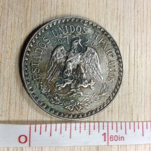 May include: Silver Mexican coin with the words "Estados Unidos Mexicanos" and a depiction of an eagle with a snake in its talons.