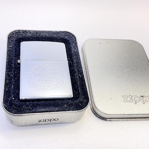 Puede incluir: Un encendedor Zippo plateado con un dise&ntilde;o grabado, presentado en una caja de metal. El encendedor presenta el texto "Terra Mater" y otras letras. La caja est&aacute; abierta, revelando el encendedor en terciopelo negro.