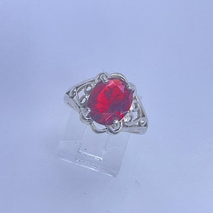May include: A silver ring with an oval, bright red gemstone. The ring has an ornate, openwork design around the gemstone. The ring is displayed on a clear, angled stand against a light background.