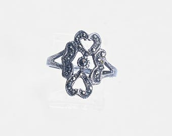 May include: A silver-toned ring with an openwork design featuring heart shapes and small, dark accents. The ring has a central, circular dark stone and is set against a white background. The ring's design is symmetrical and ornate.