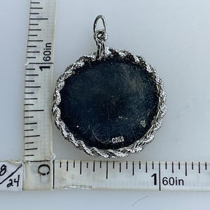 May include: Silver pendant with a round, black, textured surface and a silver rope-like border. The back of the pendant has the text "CREA GISTER" engraved on it.