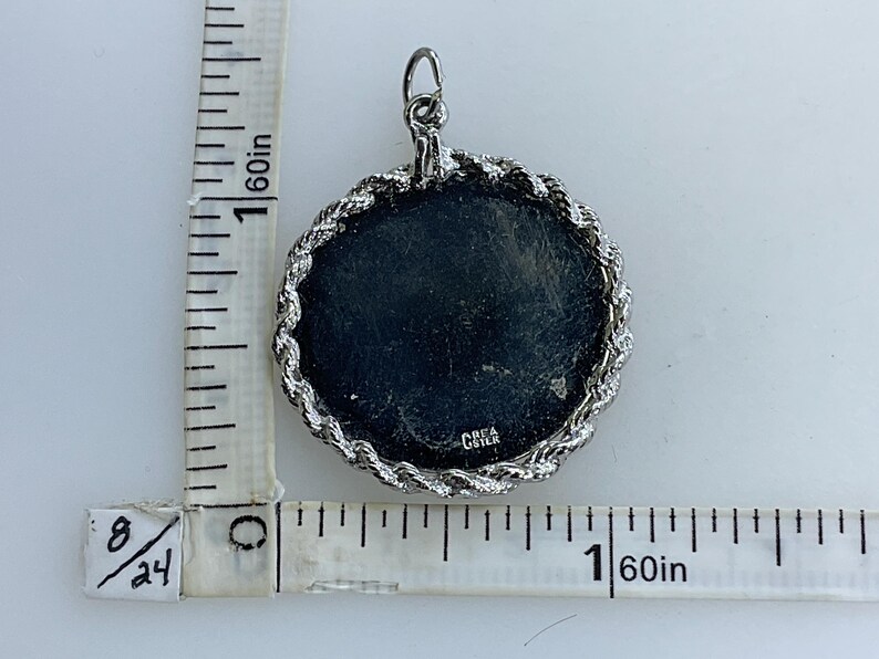 May include: Silver pendant with a round, black, textured surface and a silver rope-like border. The back of the pendant has the text "CREA GISTER" engraved on it.