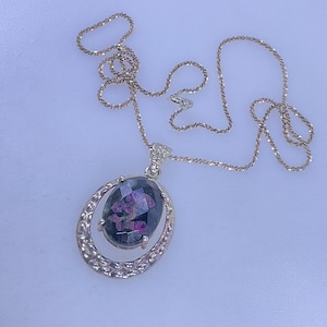 May include: A silver-toned necklace with an oval pendant. The pendant features a dark, possibly gemstone, center with pink and purple inclusions, set within a decorative, openwork frame. The chain is delicate and hangs on a white surface.