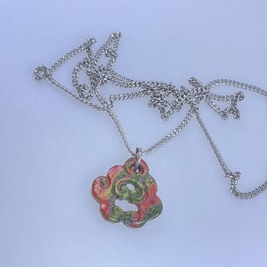 May include: A silver chain necklace with a pendant. The pendant is a cloud-like shape with a swirl design. The pendant is painted with red, yellow, and green colors.