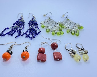 Può includere: Una collezione di sei paia di orecchini in vari stili. Gli orecchini includono frange di perline, perle pendenti e perline colorate nelle tonalità del blu, arancione, rosso e verde. Gli orecchini sono esposti su una superficie bianca.
