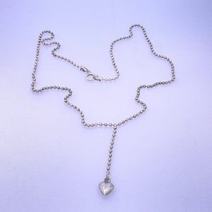 May include: A silver-toned ball chain necklace featuring a small, smooth heart-shaped pendant suspended from the center. The necklace includes a circular spring ring clasp mechanism for securing the chain around the neck.