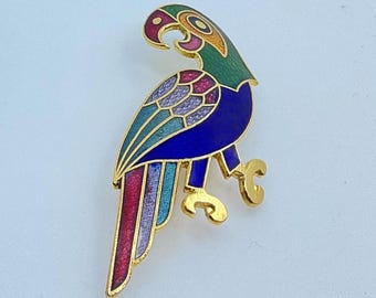 May include: A gold-toned parrot brooch with a colorful enamel design. The parrot features a green head, blue body, and wings with shades of pink, purple, and teal. The brooch has a detailed, artistic appearance.