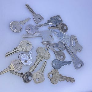 May include: A collection of various metal keys, each with unique shapes and designs. Some keys have circular or oval heads, while others have more intricate patterns. The keys are scattered on a light-colored surface.