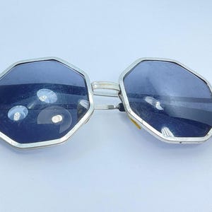 May include: Silver-framed sunglasses with dark blue lenses. The lenses have an octagonal shape. The arms are silver and black. The sunglasses are on a white surface.
