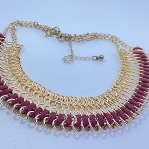 May include: Gold-tone necklace with a layered design. The necklace features a series of gold-colored chain links and burgundy-colored suede-like accents. The necklace has an adjustable chain closure.