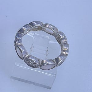 May include: A silver-toned ring with an alternating pattern of oval-shaped stones and decorative elements. The ring is displayed on a clear, angled stand against a light blue background. The stones appear to be a pale pink color.