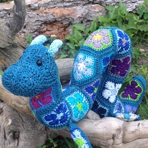 May include: A handmade, blue crochet dragon with a segmented body. The dragon features a head with small horns and a body adorned with floral patterns in shades of blue, purple, green, and white. The dragon is lying on a piece of driftwood.