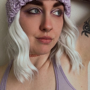 May include: A lilac crochet beanie with a dark brown square patch featuring a white X design. The beanie is made of a chunky yarn. The person is wearing a lilac tank top and has white hair.