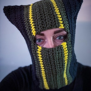 Könnte beinhalten: Handgefertigte Häkel-Balaclava in Grün und Schwarz, akzentuiert mit leuchtend gelben Streifen. Die Balaclava hat spitz zulaufende, ohrenartige Verlängerungen oben und eine Gesichtsöffnung. Die Person trägt ein schwarzes Oberteil.