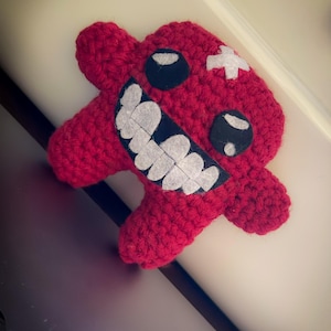 Può includere: Un mostro rosso fatto a mano all'uncinetto con occhi neri, una bocca in feltro con denti bianchi e una toppa bianca a forma di croce. Il peluche ha quattro appendici e poggia su una superficie chiara.