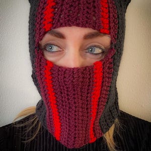 Könnte beinhalten: Handgefertigte Häkel-Balaclava in den Farbtönen Dunkelgrau, Burgunderrot und Rot. Die Balaclava hat ohrenartige Vorsprünge und rote Streifen in der Mitte. Die Gesichtsöffnung ist sichtbar.