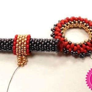 May include: A beaded jewellery piece with a red, gold, and black colour scheme. The piece is made up of a long, straight section and a circular section. The circular section is attached to the straight section. The piece is made of small beads that are strung together on a wire.