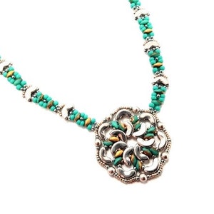 Könnte beinhalten: Eine türkisfarbene und silberne Perlenkette mit einem großen, kunstvollen Anhänger. Der Anhänger besteht aus Silber- und Türkissperlen und hat ein geometrisches Design.