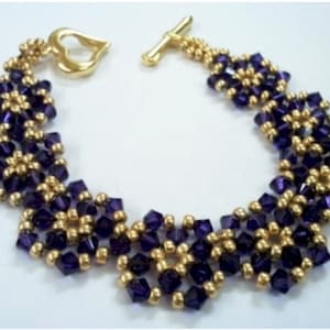 Może przedstawiać: Bransoletka z koralików złotych i fioletowych z zapięciem w kształcie serca. Bransoletka wykonana jest z małych, okrągłych koralików i charakteryzuje się powtarzającym się wzorem fioletowych i złotych koralików.