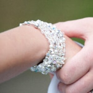 Könnte beinhalten: Ein Stapel silberner und durchsichtiger Kristallperlenarmbänder. Die Armbänder sind übereinander am Handgelenk einer Person gelegt.