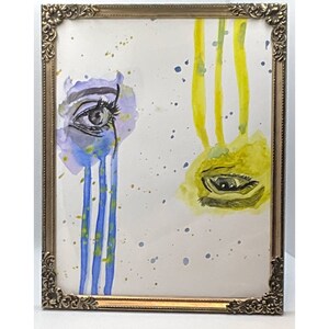 May include: A framed watercolor painting of two eyes, one with blue and yellow watercolor and the other with yellow watercolor. The eyes are painted on a white background with blue and yellow splatters.